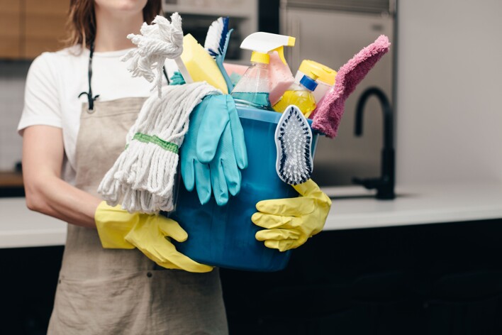 A domestic cleaner is holding a bucket full of cleaning supplies.